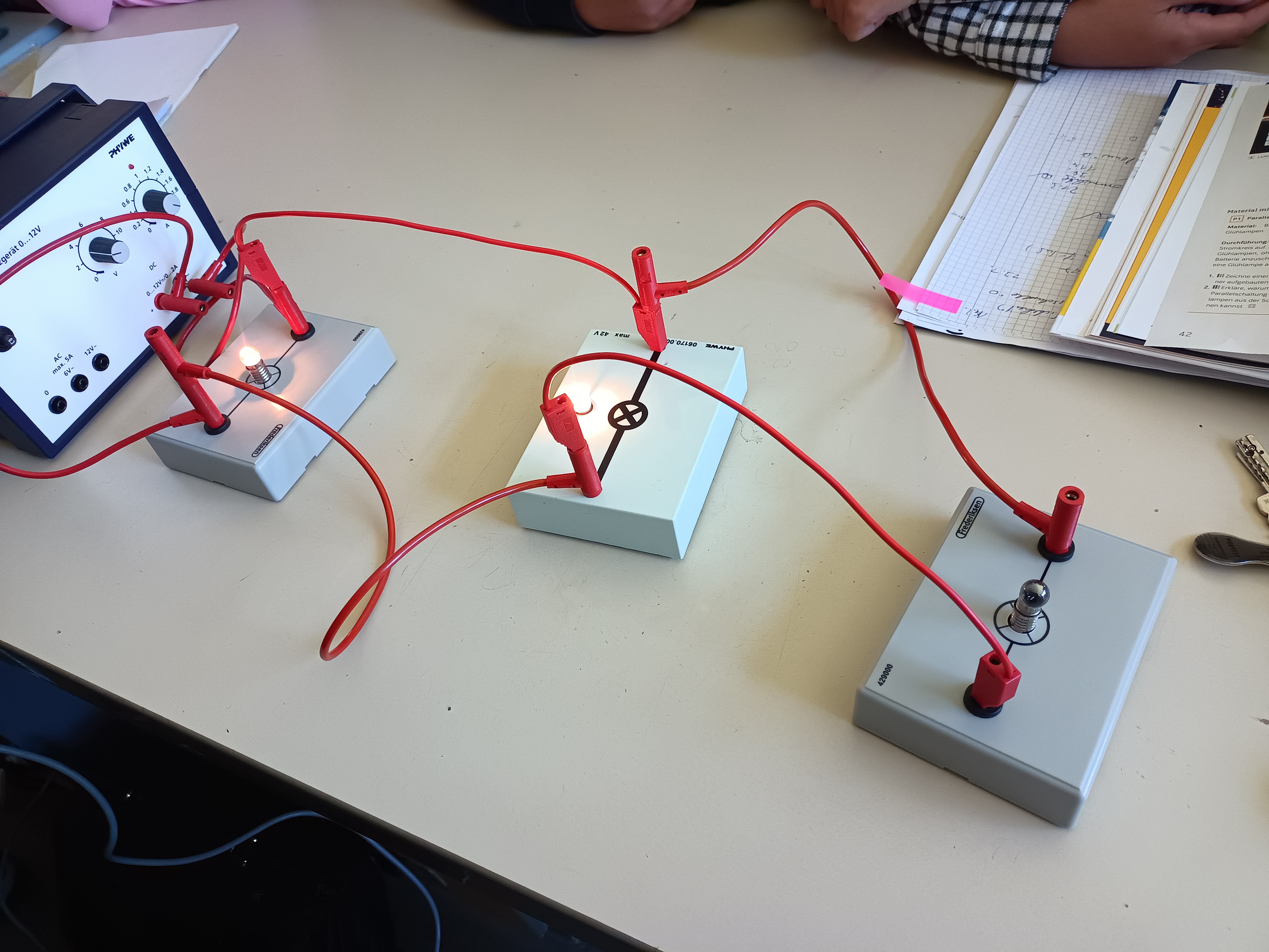 Aufgebauter Stromkreis-Versuch mit Netzgerät, Schalter und Glühlampen, verbunden mit roten Kabeln, Physikunterricht Stufe 5.